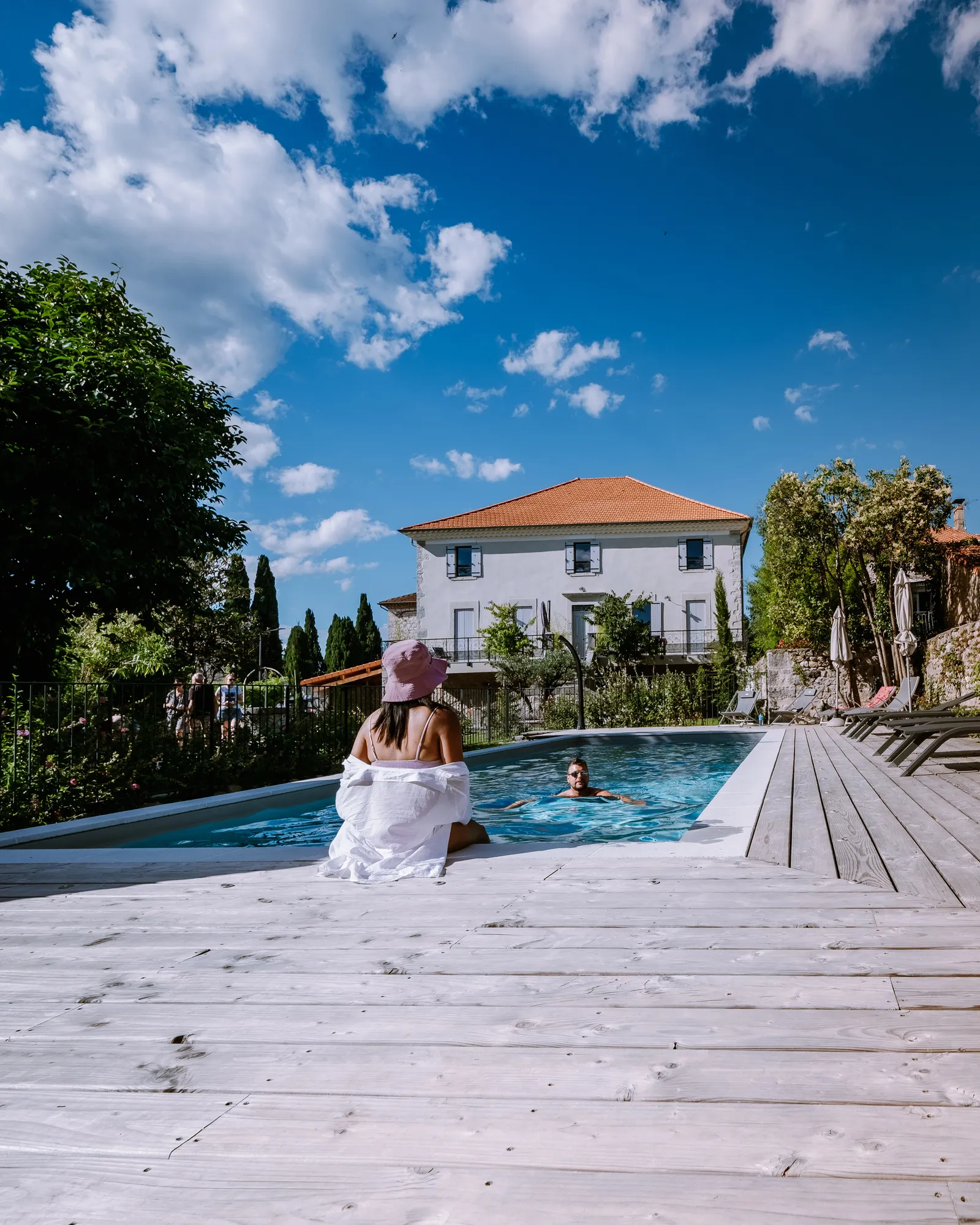 A woman in a sun hat sits at the edge of a villa swimming pool in France